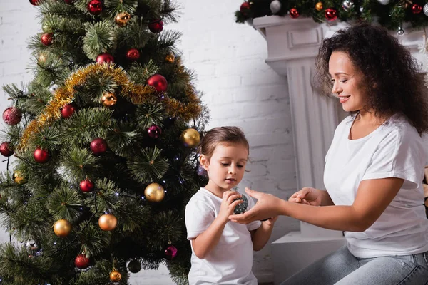 Madre e hija con bolas de Navidad sentadas cerca de pino decorado en casa - foto de stock