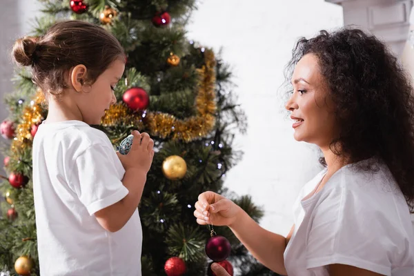 Side view of mother and daughter with christmas ball near pine at home — Stock Photo