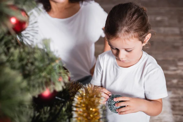 Selective focus of girl standing near mother and holding bauble near pine — Stock Photo