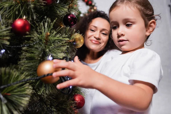 Низький кут зору дівчини, що торкається різдвяного м'яча на сосну біля матері вдома — стокове фото