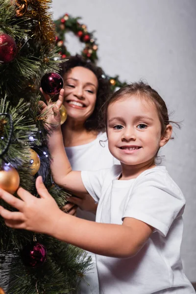 Селективный фокус девушки, трогающей рождественские балы на сосне рядом с матерью дома — стоковое фото