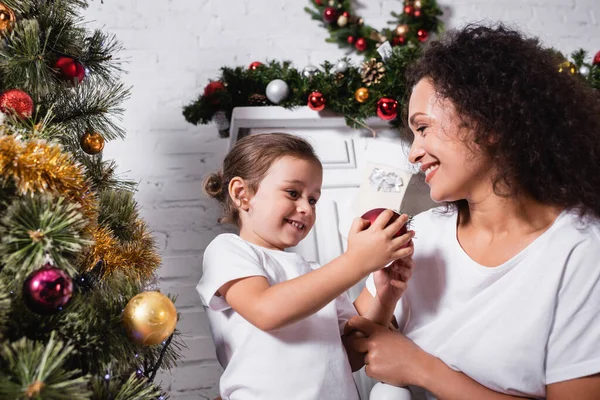 Mutter und Mädchen mit Weihnachtskugel bei geschmückter Kiefer zu Hause — Stockfoto