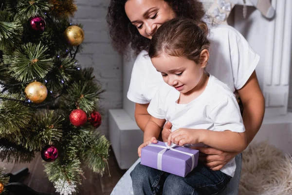 Mère avec fille ouvrant boîte cadeau près de pin festif à la maison — Photo de stock