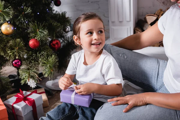 Fille avec boîte cadeau assis près de la mère et regardant loin près de pin festif — Photo de stock