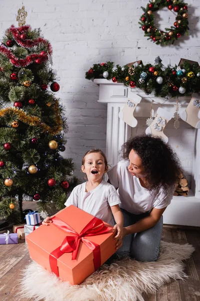 Ragazza con la bocca aperta che tiene grande scatola regalo vicino alla madre e pino festivo a casa — Foto stock