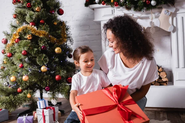 Madre con hija sosteniendo gran caja de regalo cerca de pino festivo y chimenea - foto de stock