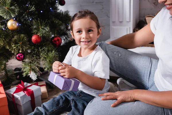 Bambina con scatola regalo guardando lontano vicino alla madre e pino decorato a casa — Foto stock