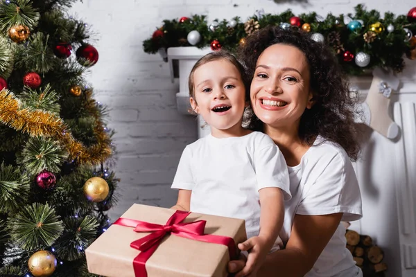 Mother hugging daughter with gift box and looking at camera near pine at home — Stock Photo