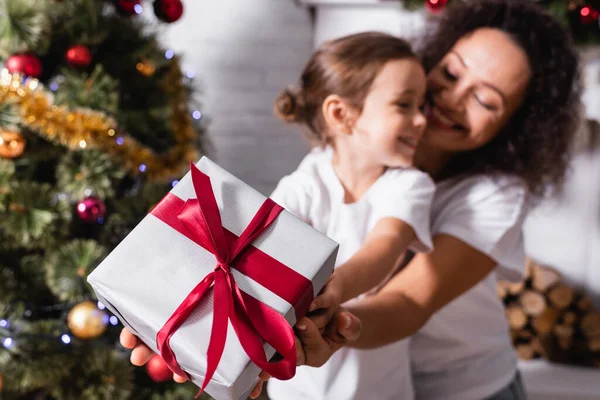 Enfoque selectivo de la madre abrazando a la hija con caja de regalo cerca del pino decorado - foto de stock