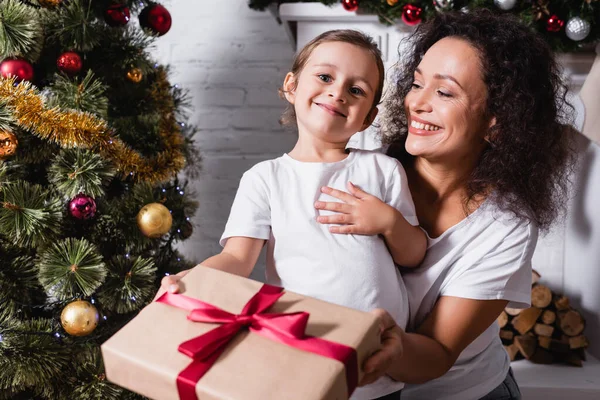 Niña con caja de regalo mirando a la cámara cerca de la madre y el pino festivo en casa - foto de stock