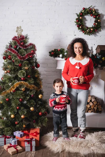 Mother and daughter with cups standing near festive pine and fireplace at home — Stock Photo