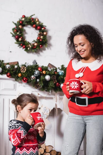 Little girl drinking from cup, standing with mother near decorated fireplace — Stock Photo