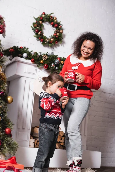 Girl drinking from cup standing with mother near decorated fireplace at home — Stock Photo