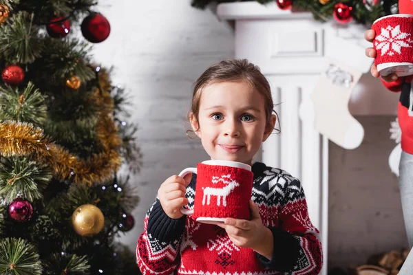 Front view of girl holding cup with knitted holder standing near festive pine — Stock Photo