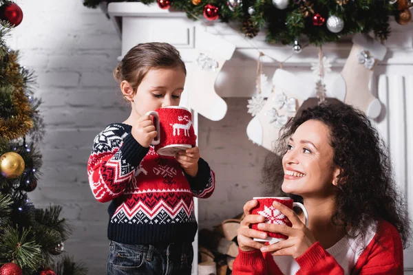 Daughter drinking from cup and standing near mother with fireplace on background — Stock Photo