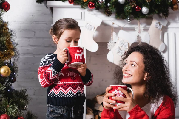 Hija bebiendo de la taza y de pie cerca de la madre con chimenea en el fondo - foto de stock