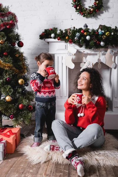 Mother sitting on rug near daughter drinking from cup near festive pine at home — Stock Photo