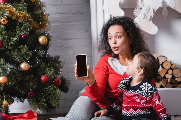 Daughter with mother showing cellphone with blank screen near pine and fireplace — Stock Photo