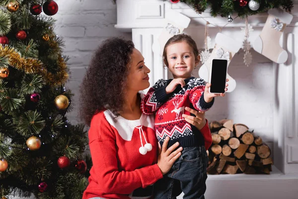 Mutter umarmt Tochter und zeigt Smartphone mit leerem Bildschirm neben Kamin — Stockfoto