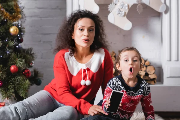 Sorpresa madre e figlia con cellulare seduto vicino a pino e camino — Foto stock