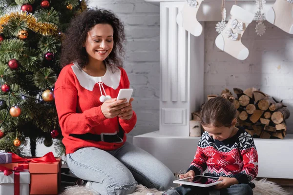 Mother and daughter using gadgets sitting near pine and fireplace at home — Stock Photo