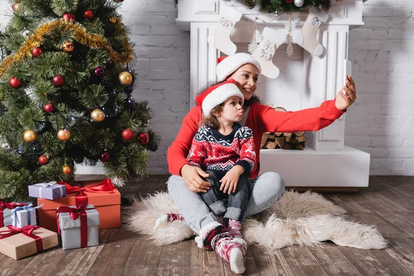 Mère et fille en chapeaux de Père Noël prenant selfie près de pin décoré à la maison — Photo de stock