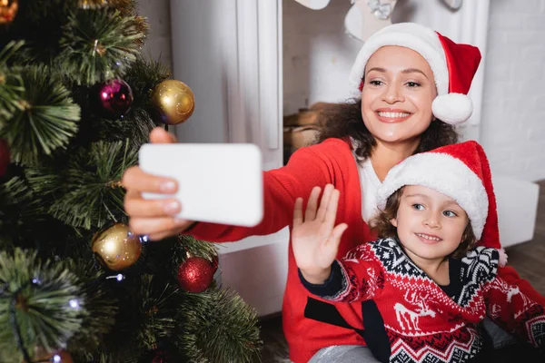 Madre e hija con la mano agitada tomando selfie cerca de pino decorado - foto de stock