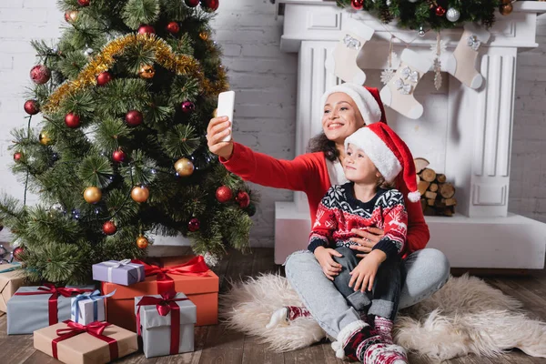 Madre e figlia in cappelli di Babbo Natale prendere selfie vicino pino festivo a casa — Foto stock