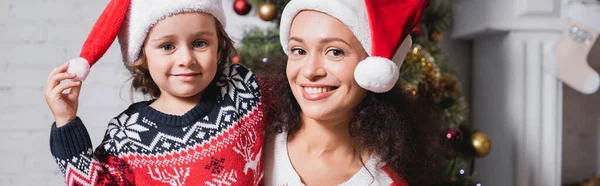 Panoramaaufnahme von Mutter und Tochter in Weihnachtsmannmützen, die in die Kamera schauen — Stockfoto