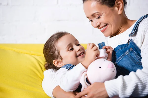 Mãe abraçando filha com brinquedo macio no sofá amarelo — Fotografia de Stock