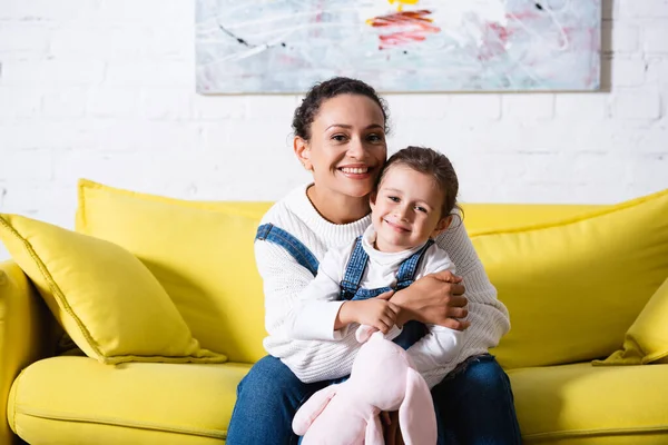Mamá sentada en el sofá y abrazando a su hija con un juguete suave en casa - foto de stock