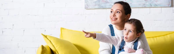 Panoramic shot of mom and daughter pointing with finger and looking away at home — Stock Photo