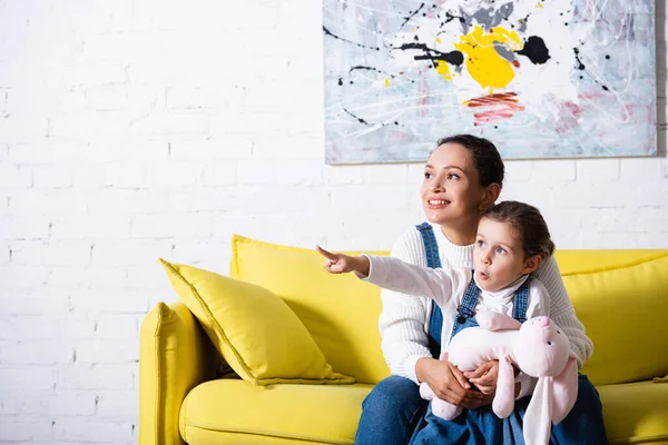 Mom hugging daughter holding soft toy and pointing with finger at home — Stock Photo