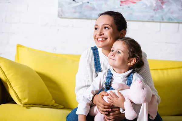 Madre abrazando a su hija y mirando hacia otro lado mientras está sentada en un sofá amarillo - foto de stock