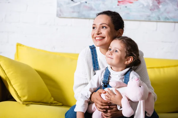 Mère étreignant sa fille et regardant loin tout en étant assis sur le canapé jaune — Photo de stock