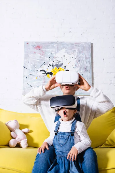 Madre e hija usando auriculares de realidad virtual sentadas en un sofá amarillo - foto de stock