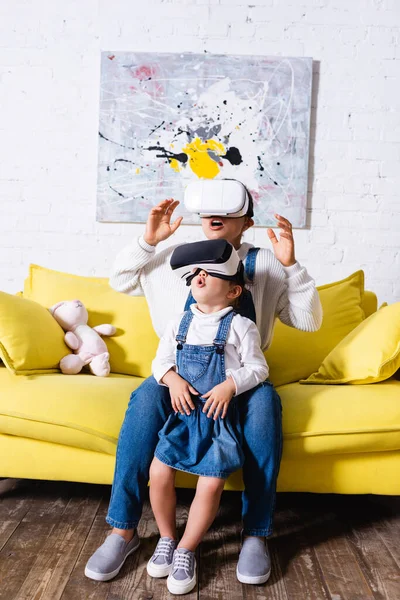 Emocionados madre e hija usando auriculares de realidad virtual cerca de sofá amarillo - foto de stock