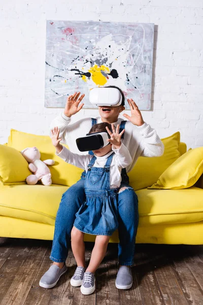 Mãe e filha com as mãos no ar usando fones de ouvido realidade virtual em casa — Fotografia de Stock