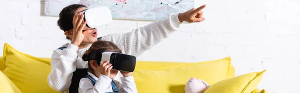 Panoramic shot of mom pointing with finger and daughter wearing VR headsets — Stock Photo