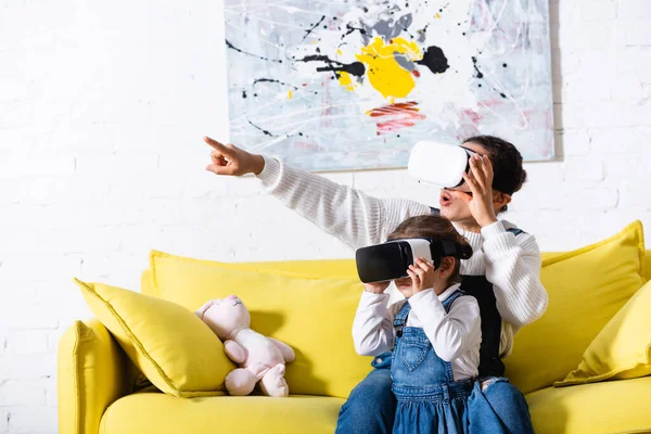 Mère pointant avec le doigt et la fille en utilisant des casques de réalité virtuelle à la maison — Photo de stock