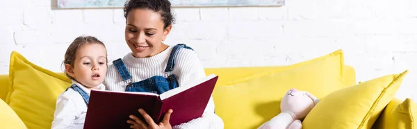 Foto panorámica de madre e hija leyendo libro en sofá amarillo en casa - foto de stock