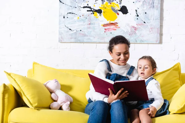 Madre e hija leyendo un libro y sentadas en un sofá amarillo en casa - foto de stock