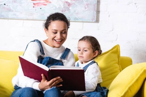 Mãe e filha lendo livro no sofá amarelo perto da foto em casa — Fotografia de Stock