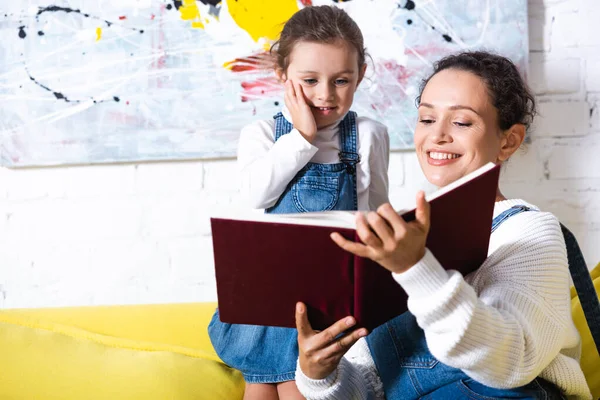 Hija de pie cerca de madre sosteniendo libro con foto en el fondo - foto de stock