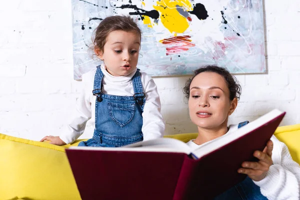 Fille excitée debout près de la mère tenant livre avec photo sur fond — Photo de stock