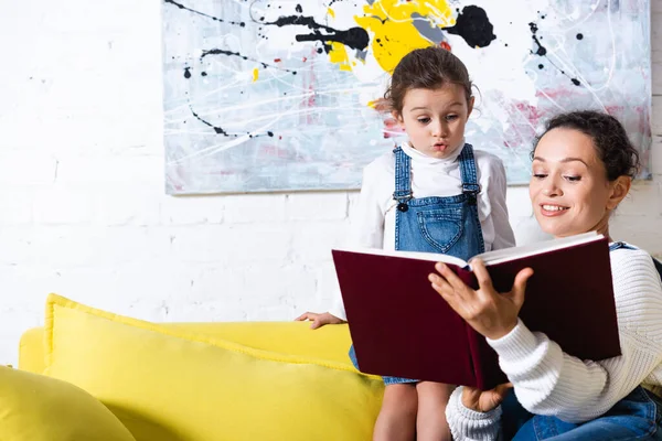 Aufgeregte Tochter steht mit Buch neben Mutter auf gelbem Sofa — Stockfoto