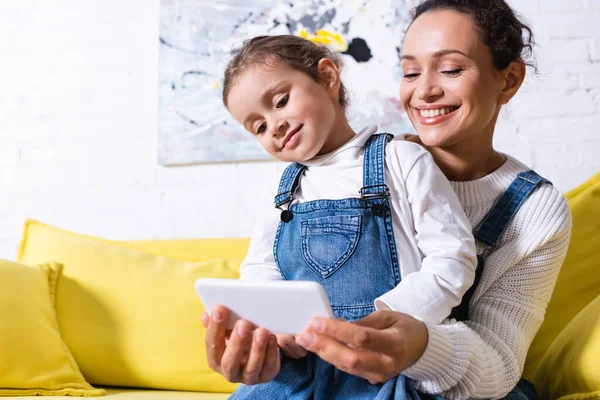 Mãe abraçando filha e tomando selfie no sofá amarelo em casa — Fotografia de Stock