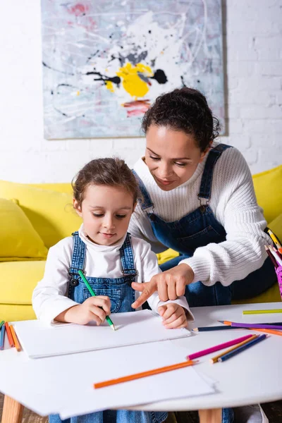 Mère pointant du doigt et fille dessinant avec des crayons de couleur à la maison — Photo de stock