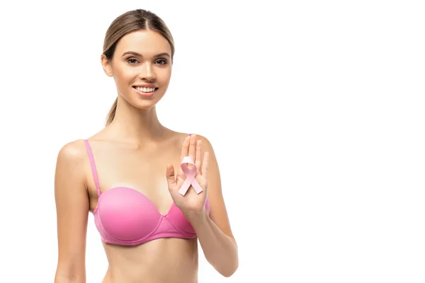 Femme brune en soutien-gorge rose regardant la caméra tout en montrant un ruban de sensibilisation au cancer du sein isolé sur blanc — Photo de stock