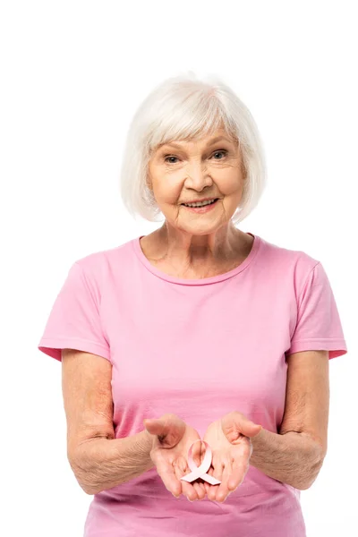 Donna anziana guardando la fotocamera mentre mostra nastro rosa di consapevolezza del cancro al seno isolato su bianco — Foto stock